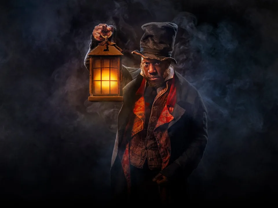 Scrooge in a battered top hat holds up a glowing lantern in the smoke, A Christmas Carol at The Old Vic