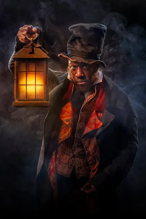 Scrooge in a battered top hat holds up a glowing lantern in the smoke, A Christmas Carol at The Old Vic