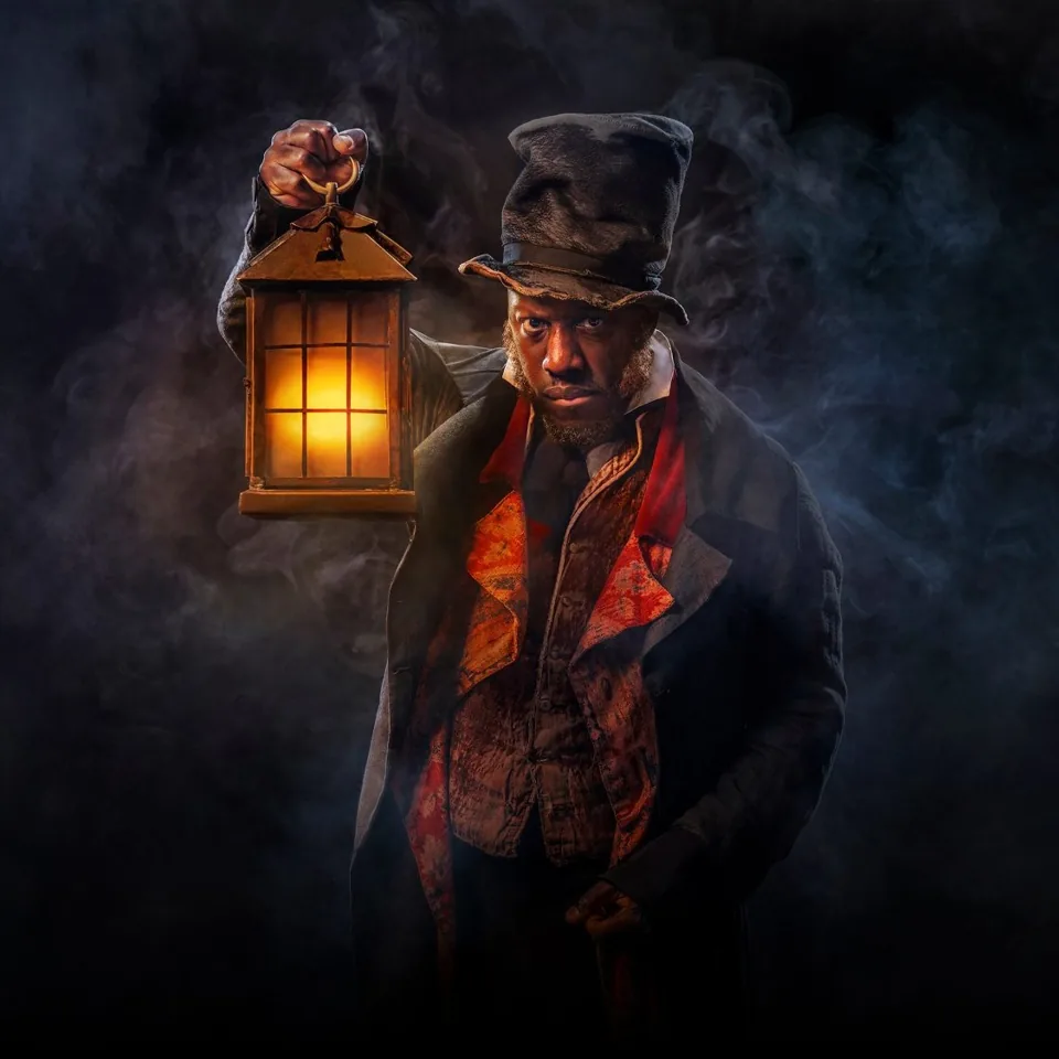 Scrooge in a battered top hat holds up a glowing lantern in the smoke, A Christmas Carol at The Old Vic