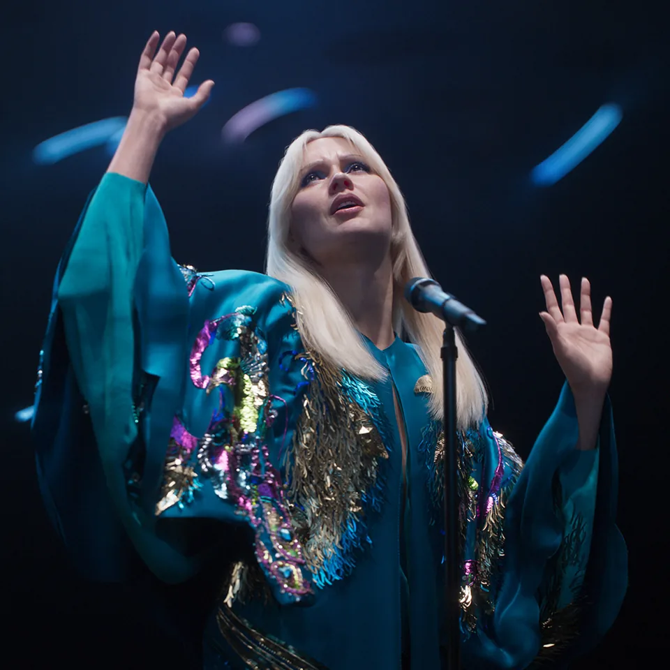 An ABBA avatar in a teal sequinned robe raises both hands, ABBA Voyage, ABBA Arena