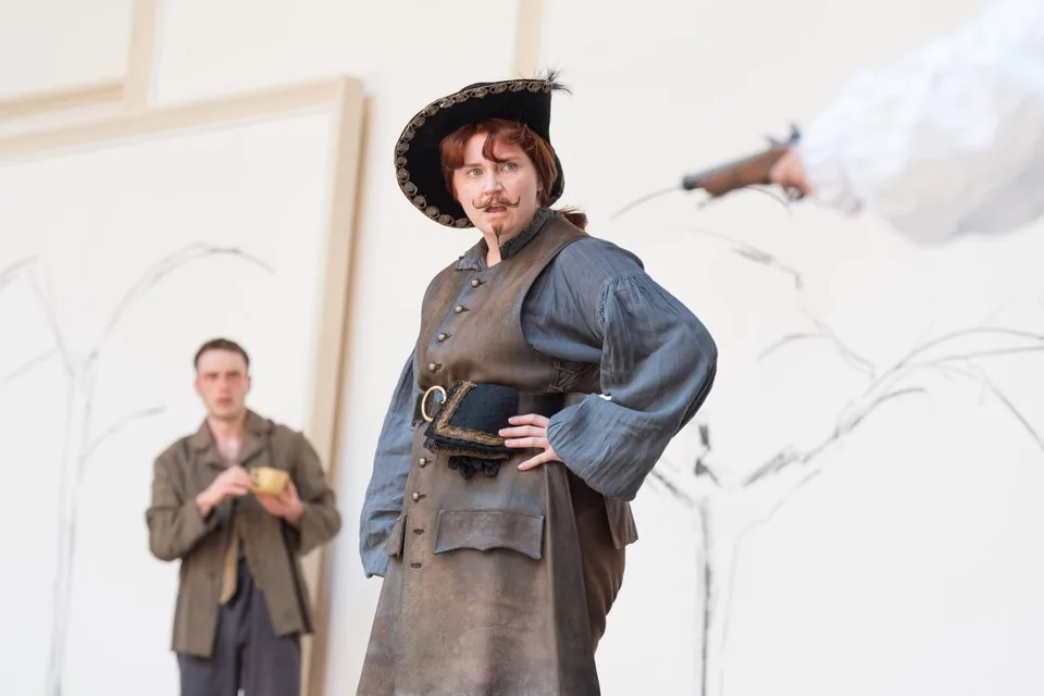 A figure in a black hat faces a gun, As You Like It, the Globe