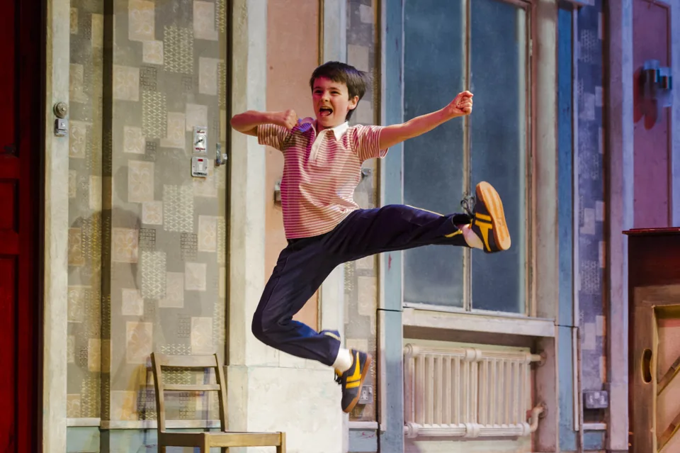 Billy leaps high with a kick by the kitchen wall, Billy Elliot the Musical