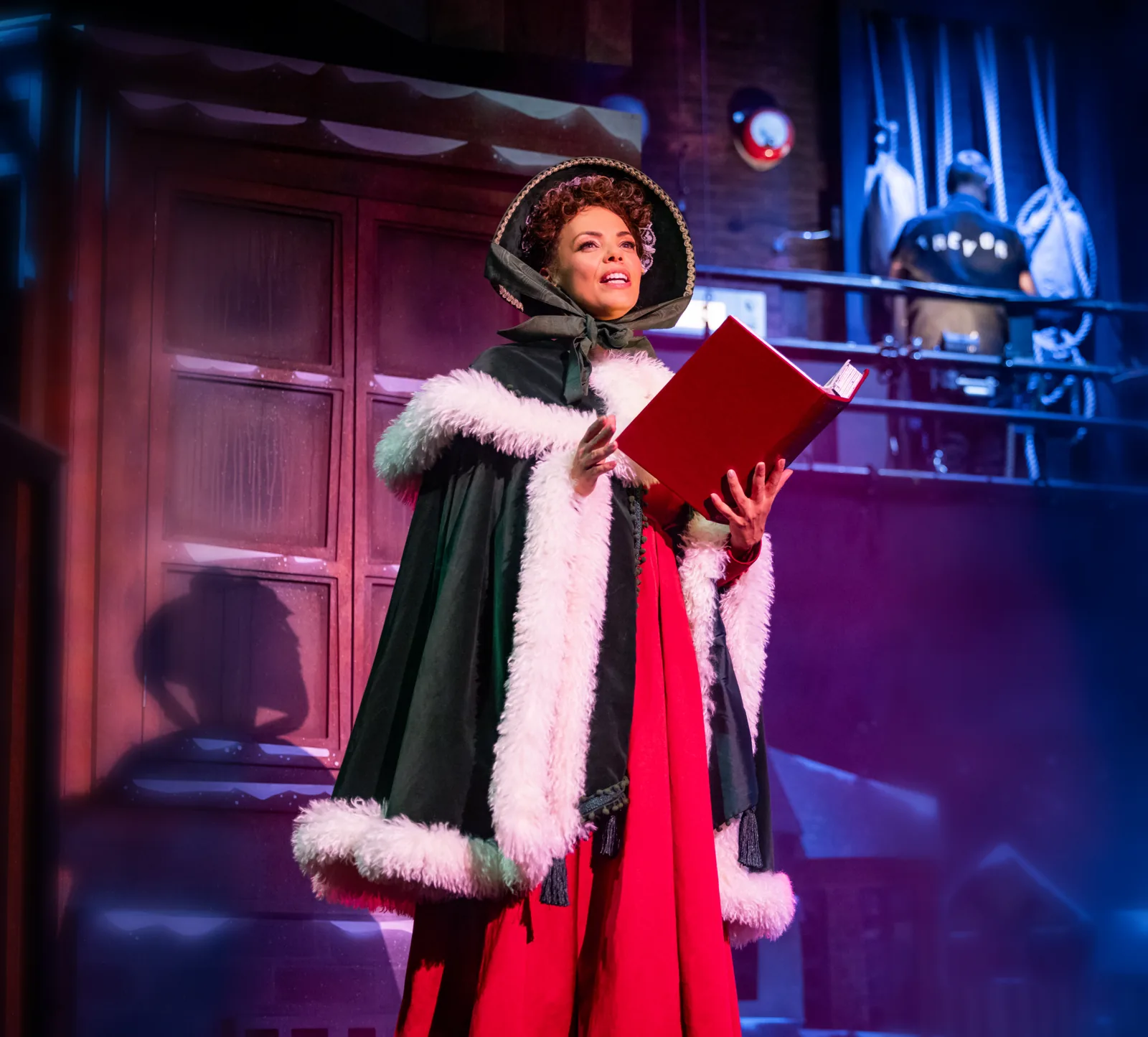 A woman in a fur-trimmed cloak reads a red book, Christmas Carol Goes Wrong, Wyndham's