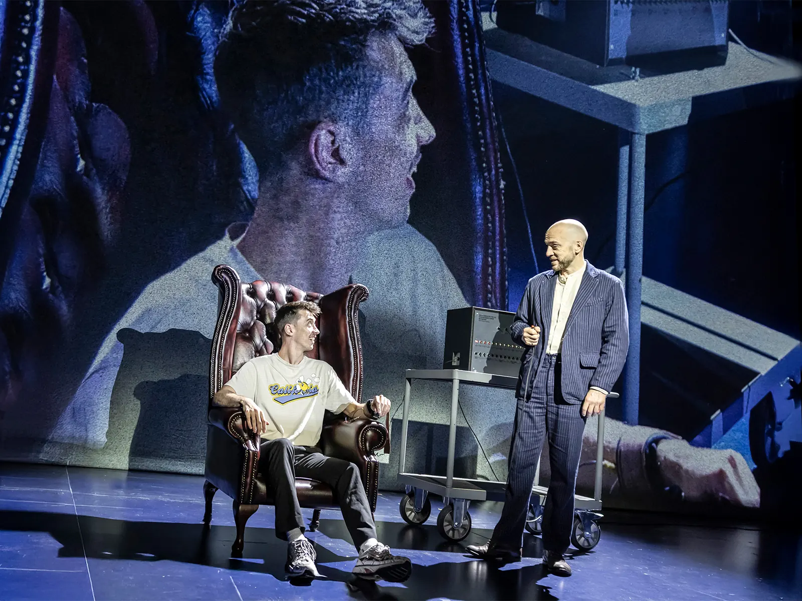A volunteer in a leather armchair as Derren Brown stands by, a close-up on the screen behind, Only Human at the Apollo Theatre
