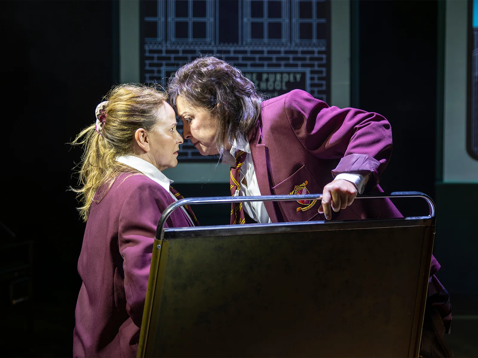 Ria Jones and Sally Ann Triplett lean close over a desk, Fourteen Again, Menier