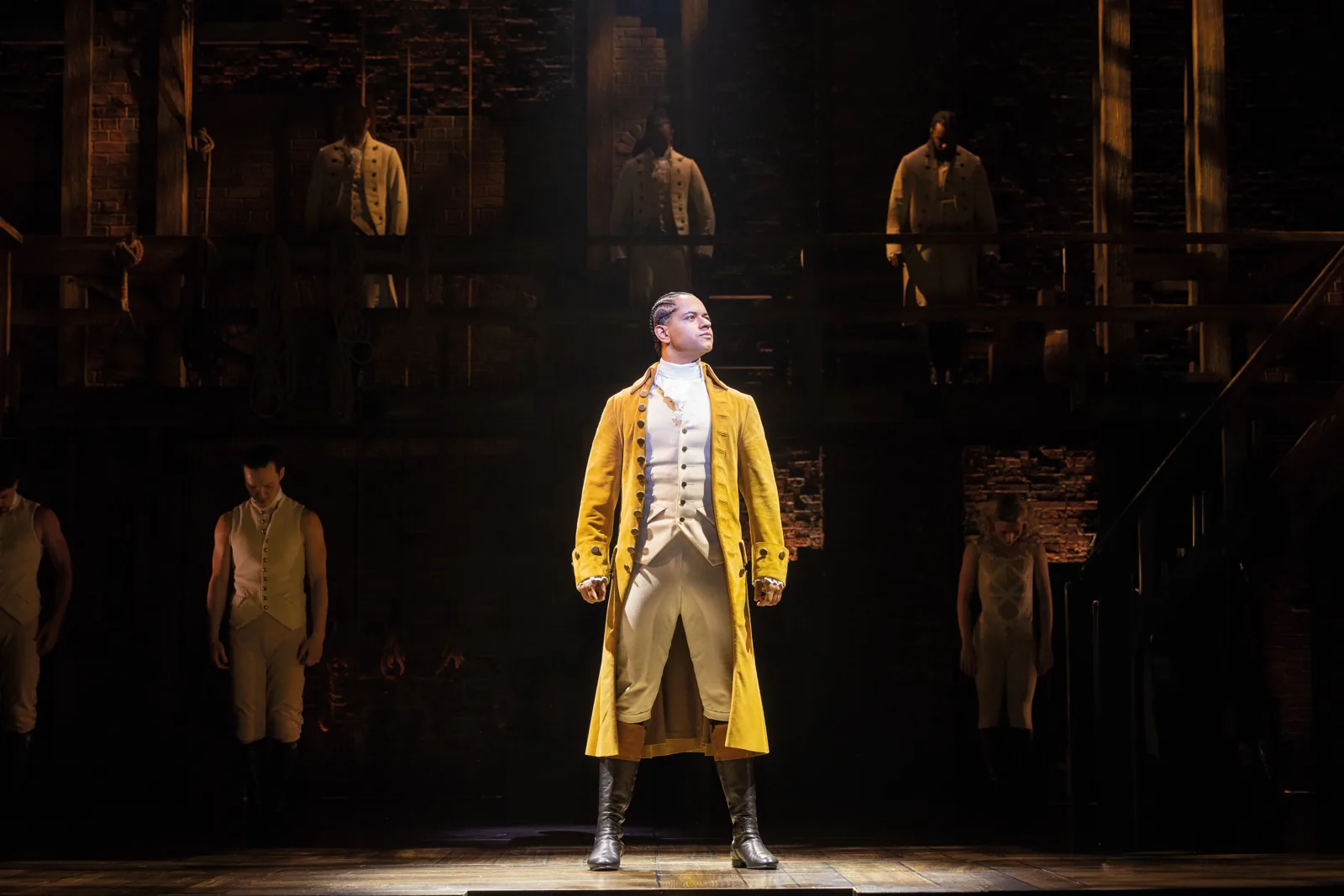 Stephenson Ardern-Sodje as Hamilton stands in a gold coat in a spotlight, Victoria Palace Theatre