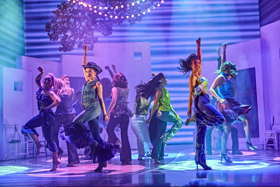 Women in flares and sequins dance under string lights in Mamma Mia!, Novello Theatre