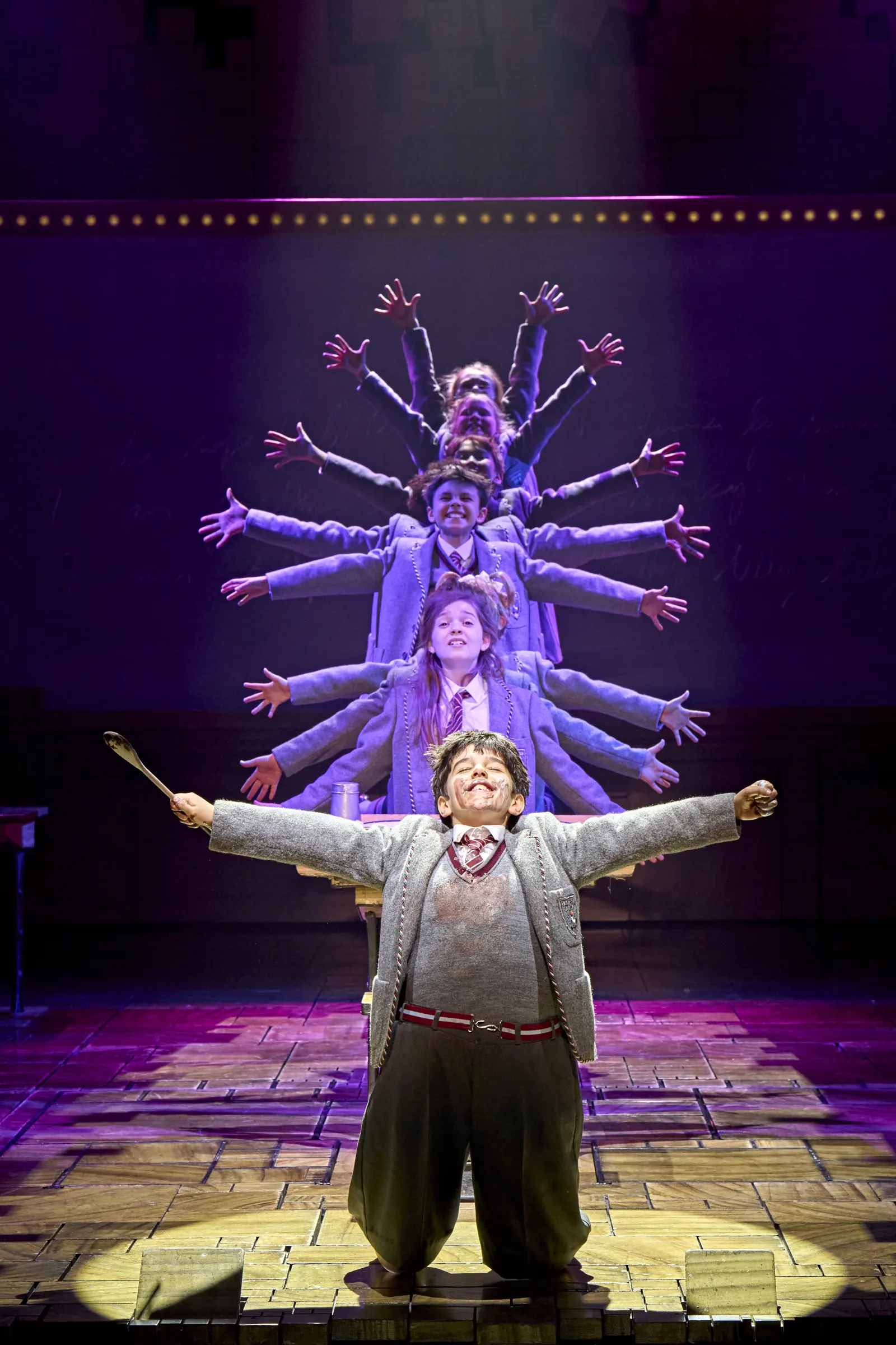 A boy flings his arms wide before fanned-out pupils, Matilda The Musical, Cambridge Theatre