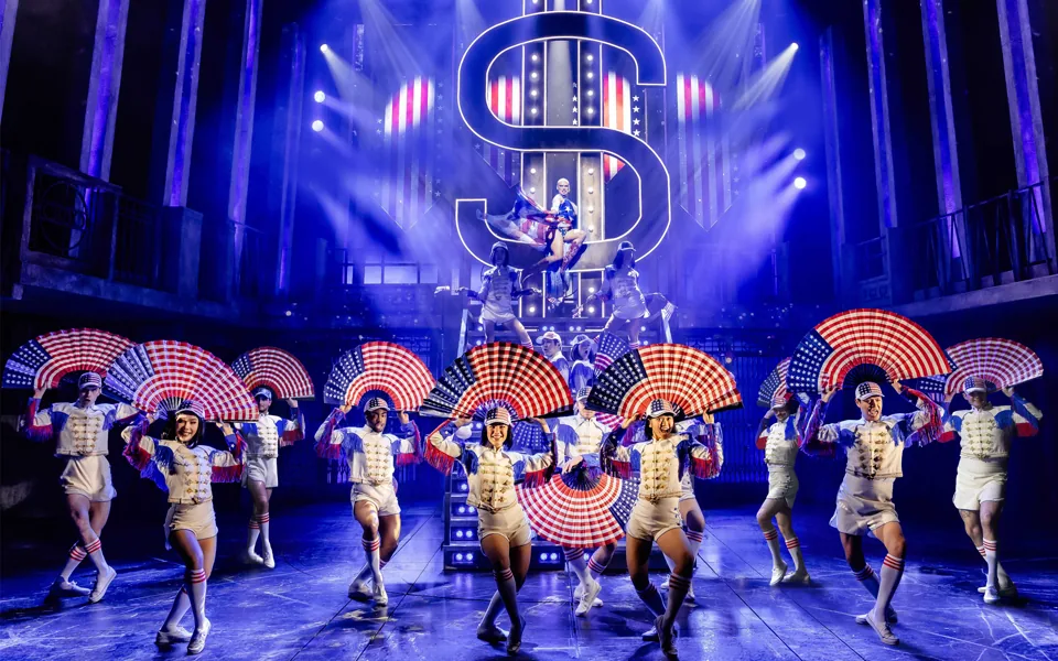 Dancers wave stars-and-stripes fans beneath a huge neon dollar sign, Miss Saigon, Prince Edward Theatre