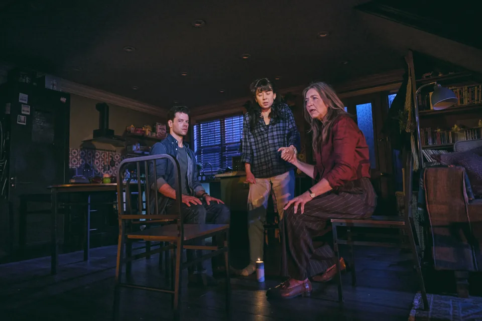 Three people talk in a dimly lit living room in Paranormal Activity, Ambassadors Theatre