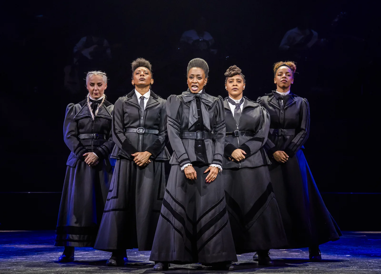 Five women in belted black coats stand in a row in Sylvia, Royal Albert Hall