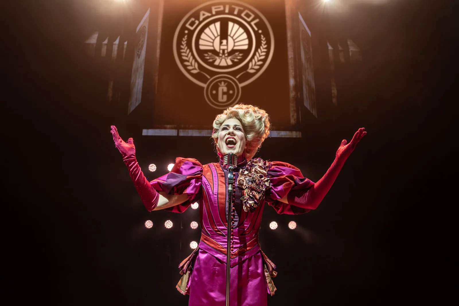 Effie Trinket, arms wide, under the Capitol crest in The Hunger Games: On Stage
