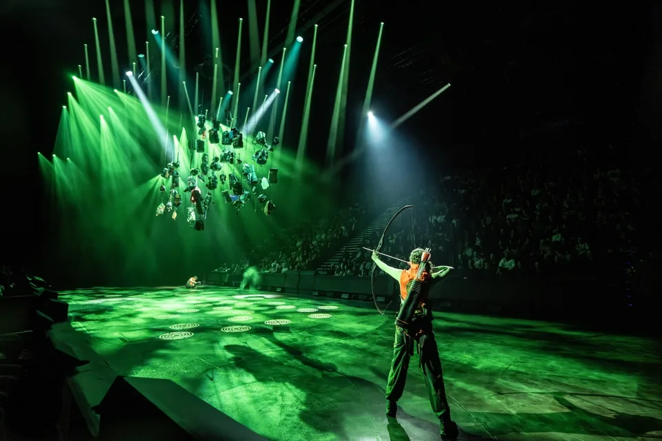 Katniss draws her bow in the green-lit arena, The Hunger Games: On Stage