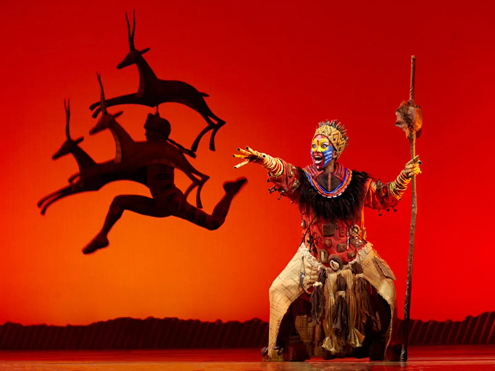 A face-painted performer with a staff as gazelles leap, The Lion King, Lyceum Theatre