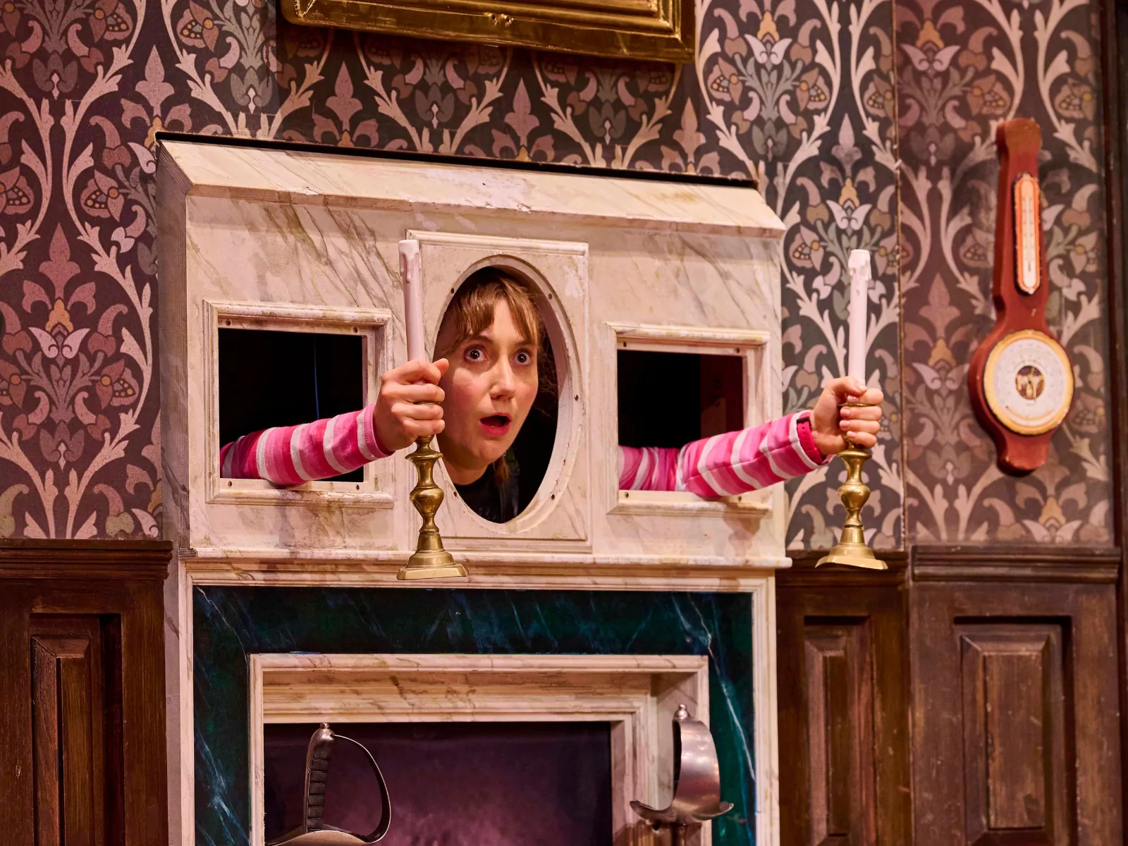 A performer peers out of the mantelpiece holding candlesticks, The Play That Goes Wrong, Duchess Theatre