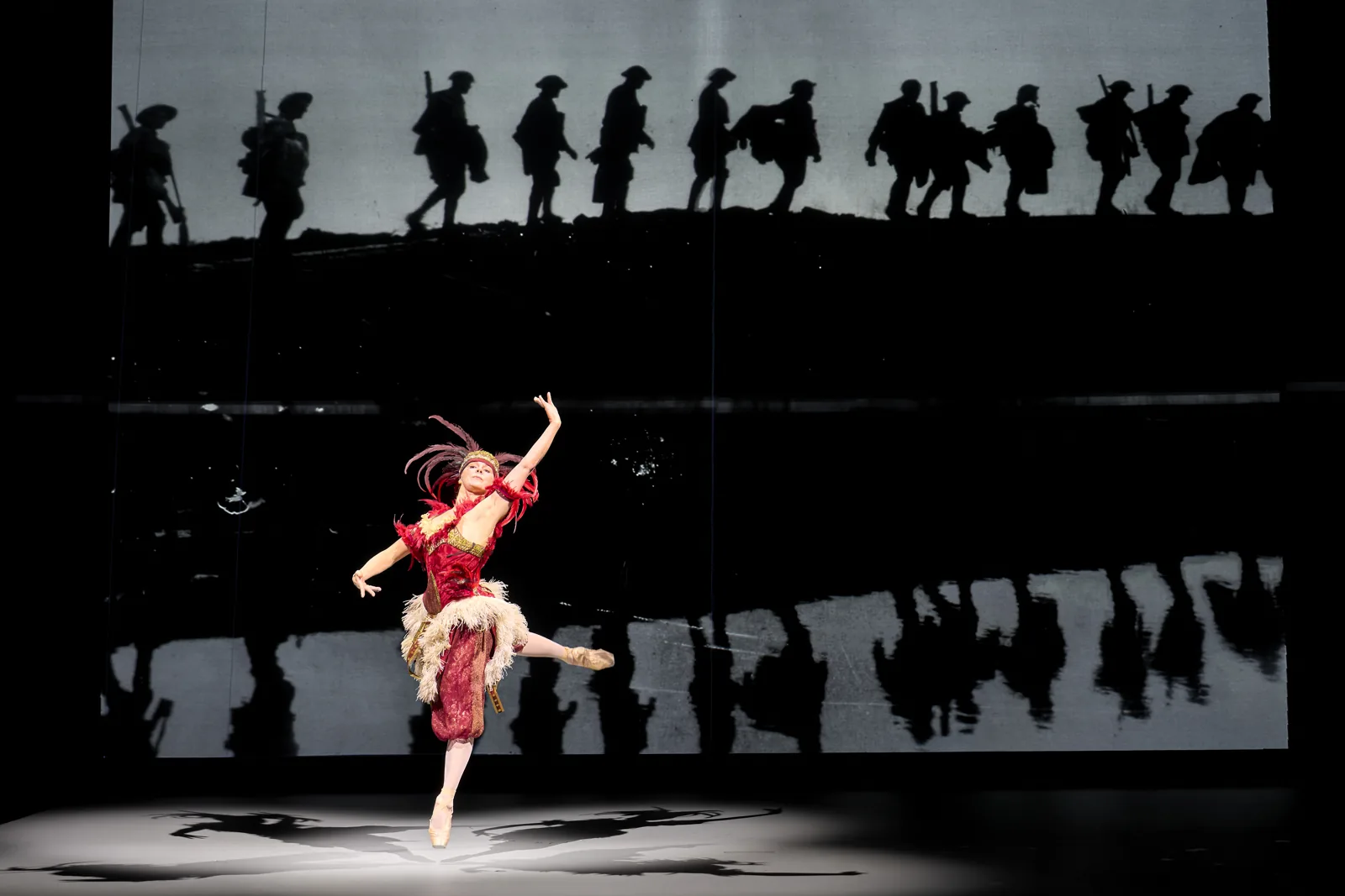 Natalia Osipova leaps in red before marching soldiers in The Standard of Living, Theatre Royal Haymarket