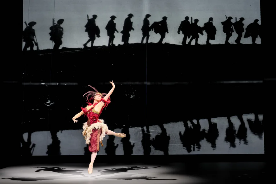 Natalia Osipova leaps in red before marching soldiers in The Standard of Living, Theatre Royal Haymarket